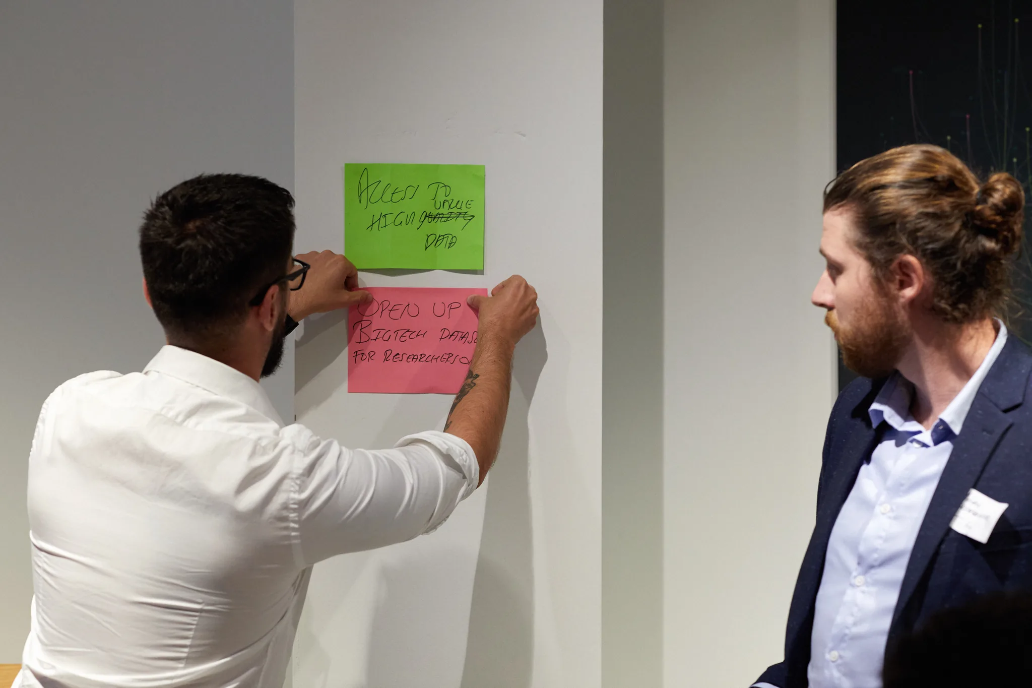 A participant placing a handwritten note reading “open up bigtech datasets for researchers” onto a wall of ideas