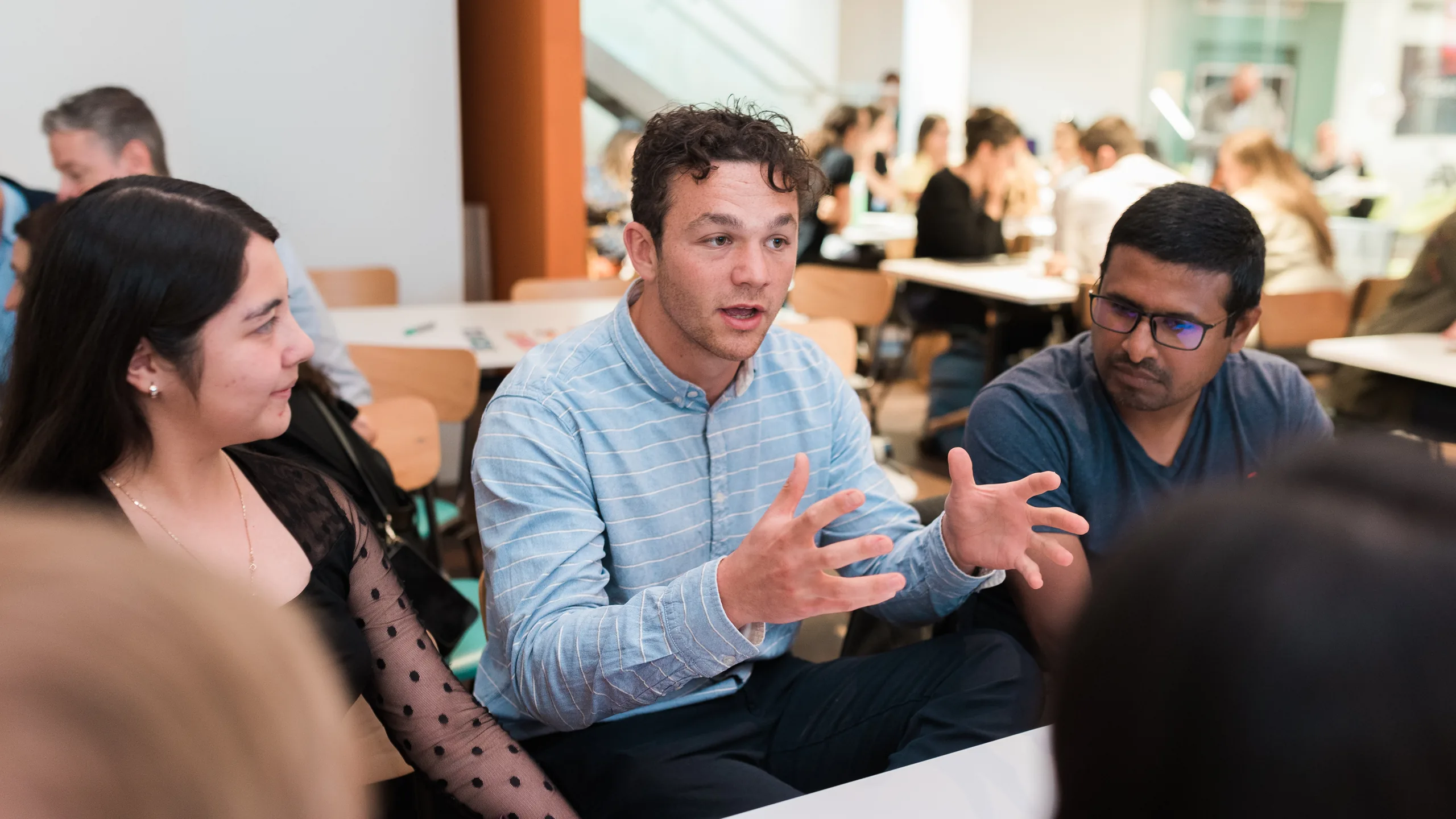 A founder explaining an idea with open hands in a daylit UCL seminar room, with other cohort tables working behind