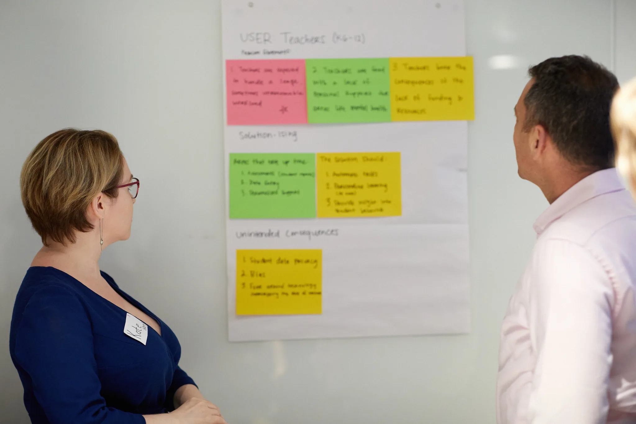 Two people reviewing a wall canvas that maps teacher problem statements, candidate solutions, and unintended consequences